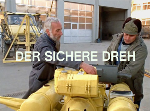 Titelbild DER SICHERE DREH mit zwei Arbeitern an einer hellgelben Kleindiesel-Baumaschine (Rüttelplatte zur Bodenverdichtung) in einem Innenhof eines Straßenbauunternehmens