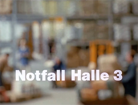 Titelbild des Kurzfilmes „Notfall Halle 3“ mit einer unscharfen Lagerhalle, Lagerarbeitern und Gabelstapler, Teil 2 aus der Erste-Hilfe-Serie "Rette mich, wer kann"