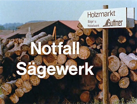 Titelbild des Kurzfilmes "Notfall Sägewerk" mit gestapelten Baumstämmen, davor ein Wegweiser mit der Aufschrift „Holzmarkt Suttner - Säge- und Hobelwerk“, Teil 6 aus der Erste-Hilfe-Serie "Rette mich, wer kann"