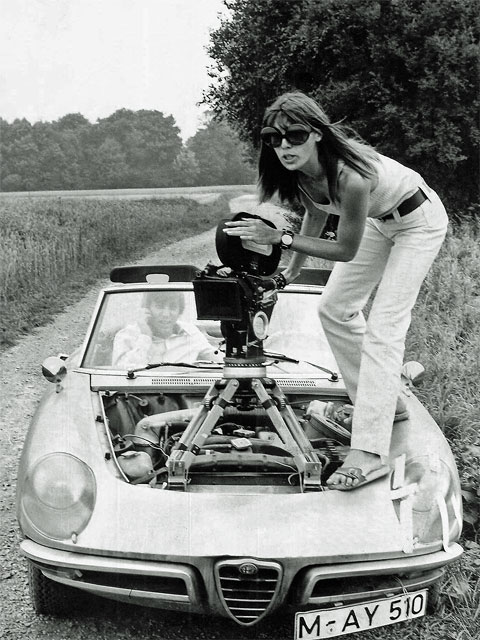 Standfoto am Waldrand von Dreharbeiten für den Kino-Film „Nicht fummeln Liebling !“ mit Regisseurin May Spils auf einem Alfa Romeo Cabrio mit offenem Verdeck stehend, auf dessen Motor eine Filmkamera montiert ist, im Cabrio sitzt Schauspieler Werner Enke