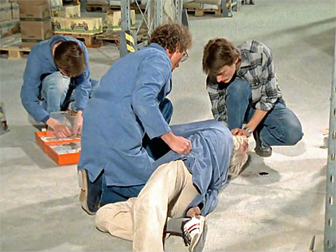 Drei Lagerarbeiter als Ersthelfer, zwei von ihnen bringen den Verletzten mit Kopfverband in die stabile Seitenlage, ein Arbeiter ordnet den orangen Verbandskasten in der Lagerhalle